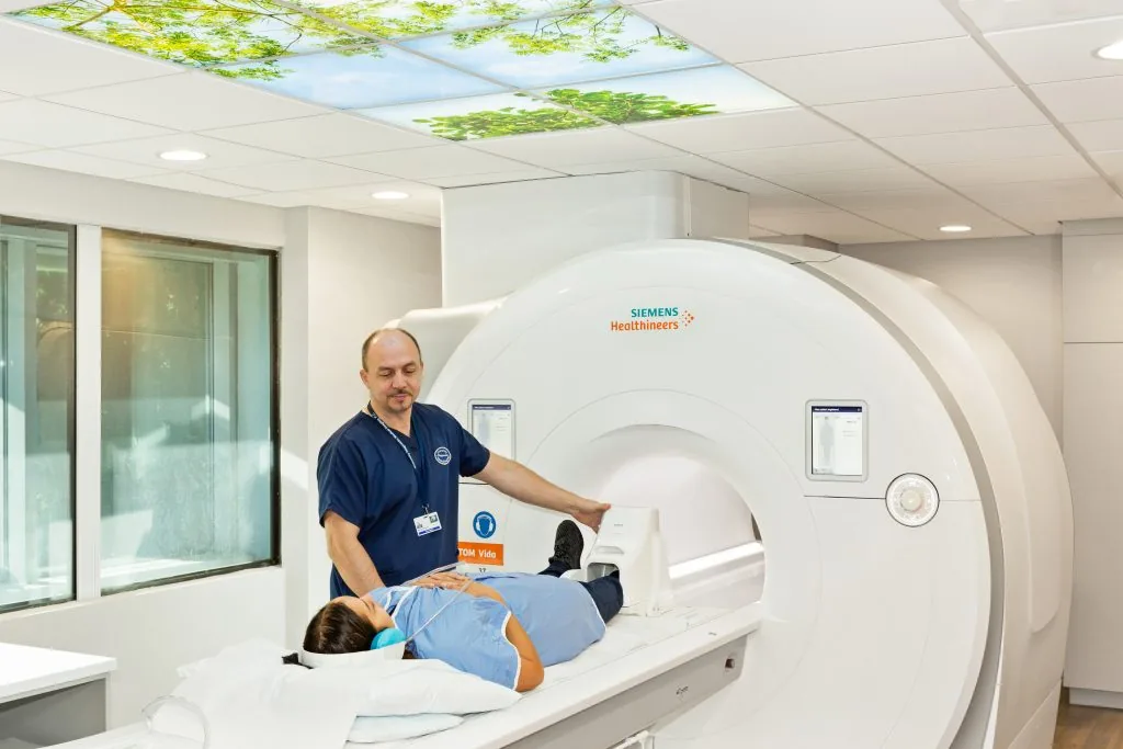 MRI technologist assisting a patient lying on the table of a 3T MRI scanner, with headphones in place